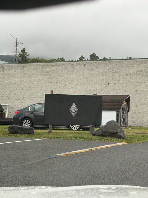A black canvas displaying a silver/diamond Ethereum logo stands prominently in a parking lot, with a car and a small structure behind it. The image ties to the post about @ethlabs_org's team offsite, with the Ethereum symbol symbolizing the team's bullish commitment to the project.