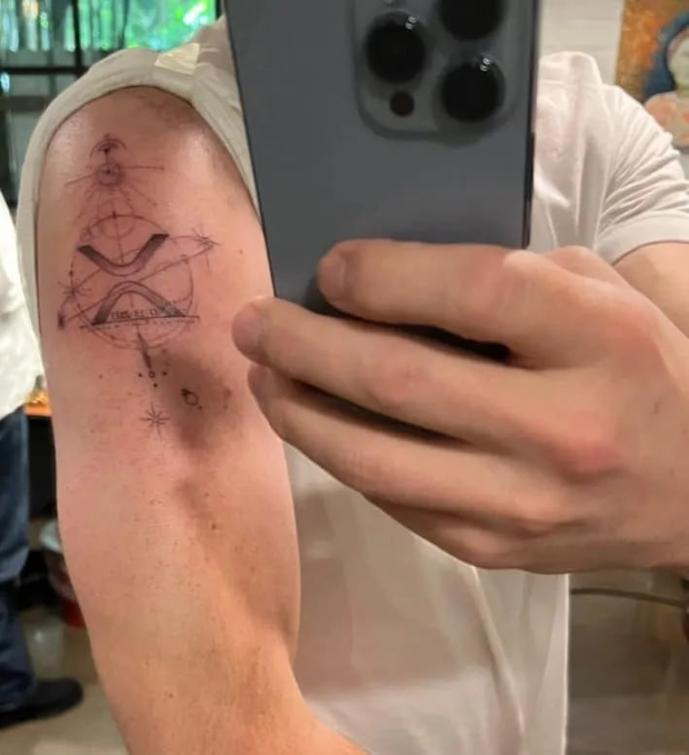 A person takes a mirror selfie showcasing a fresh tattoo on their upper arm, featuring geometric designs, constellations, planets, and the text "WE'RE ALL JUST DUST IN THE UNIVERSE," symbolizing XRP's cosmic and interconnected theme.