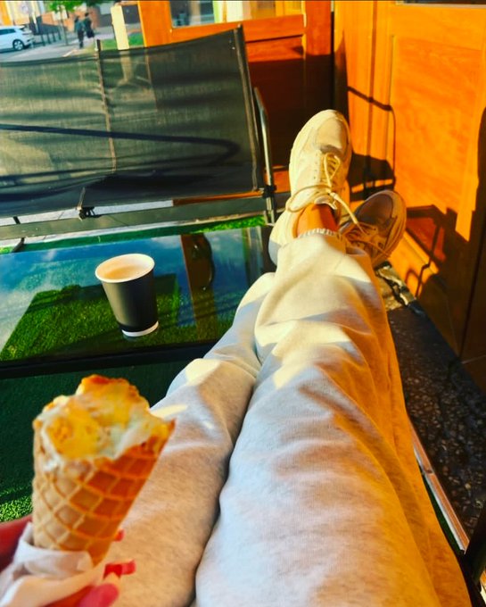 A first-person perspective photo of someone relaxing outdoors, showing their legs crossed in grey sweatpants and white sneakers, holding an ice cream cone with a coffee cup on the glass table beside them.