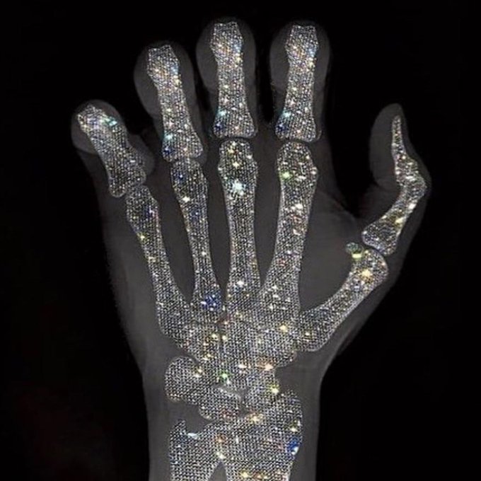 An X-ray or skeletal view of a hand heavily encrusted with sparkling diamonds or rhinestones covering the bones against a black background.
