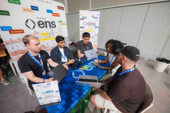 Judges from @ensdomains are seen here evaluating projects at ETHGlobal Lisbon, with the event offering $5,000 in prizes for builds that utilize ENS names, text records, and cross-chain resolvers. The image directly depicts the judging session described in the post, with participants seated around a table reviewing content on laptops.