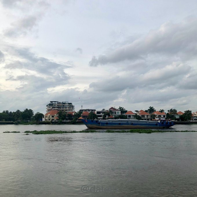 A blue cargo boat travels along a river under a cloudy sky, with residential buildings and palm trees lining the distant shore.