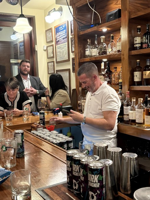 A photo taken inside a bar showing a man in a white shirt looking at his phone behind the counter, with shelves of liquor bottles behind him, customers in the background, and several cans of 'KIRIN' beer visible in the foreground.