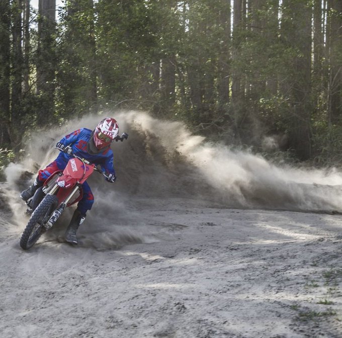 A rider in red and blue gear executes a sharp turn on a dirt bike, kicking up a dramatic cloud of dust through a forested trail. The image embodies the journey-focused message, highlighting the thrill and skill-building experience of off-road riding rather than simply reaching an endpoint.