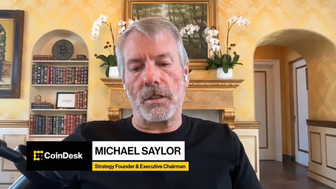 Michael Saylor, Strategy Founder & Executive Chairman, speaking in a CoinDesk interview segment while seated in a room with built-in bookshelves and orchids.
