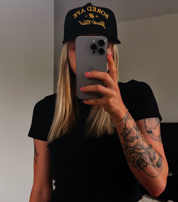 A mirror selfie of a blonde woman wearing a black shirt and a black 'BORN TO RAGE' cap, holding an iPhone, with a floral tattoo sleeve on her arm.