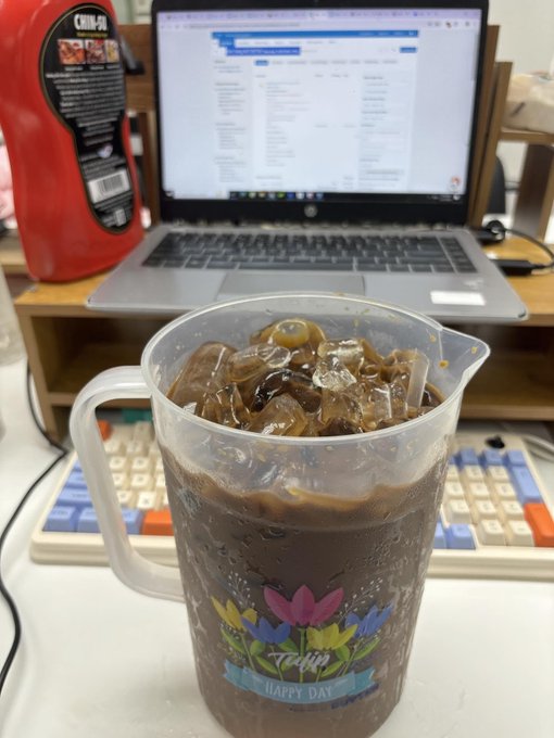 A plastic pitcher filled with iced coffee sitting on a desk in front of an HP laptop displaying a webpage, with a red dish soap bottle visible to the left.