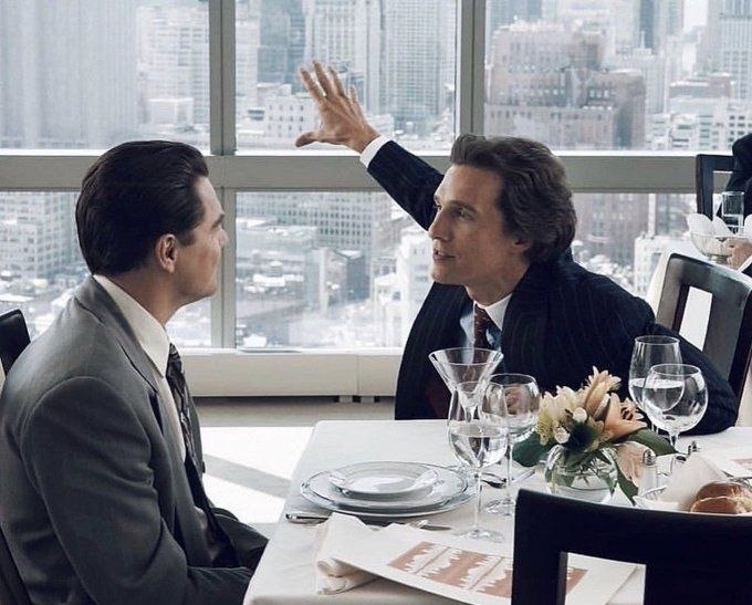 Two men in suits dine at a restaurant table with a city view, the man on the right gesturing emphatically while speaking. The image conveys the "controlled environment" and human negotiation aspect, visually mirroring the post's theme of prediction markets being subject to behind-the-scenes power dynamics and legal disputes rather than truly open platforms.