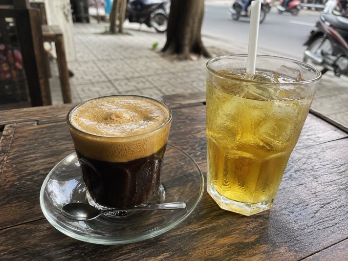 A Vietnamese iced coffee with condensed milk sits beside a glass of iced green tea on a wooden sidewalk table, capturing a refreshing morning coffee break that contrasts with the post's discussion about upcoming changes in a "bkt" policy mechanism.