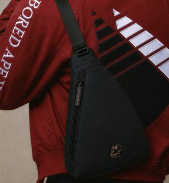 A person wearing a red jacket with 'BORED APR' visible is carrying a black triangular crossbody bag featuring a zipper and a small logo on it.