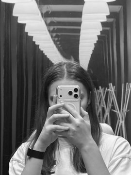 A black-and-white mirror selfie of a young woman with long hair holding a smartphone that covers her face, taken in what appears to be a modern hallway or corridor with overhead lights.