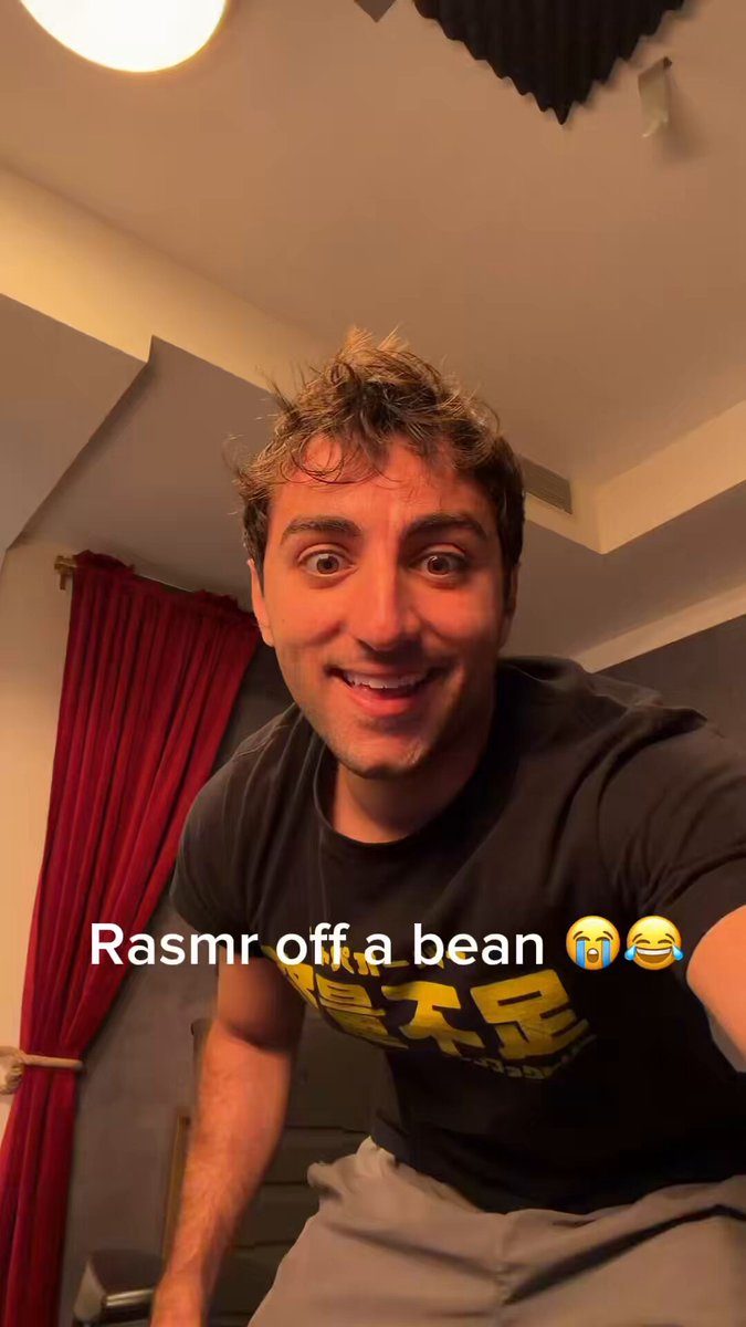 The image features a man wearing a black t-shirt with yellow text, which reads "Rasmr off a bean". The man has short hair and is smiling at the camera. There are also two emojis present in the image - one of a laughing face and another of a crying face. The background shows a red curtain and a white wall, creating an interesting contrast with the man's attire.