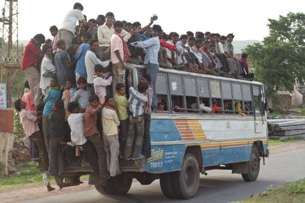 An overcrowded bus in India packed with passengers sitting on the roof and hanging off the back, with 'India TRAVELS' visible on the side.