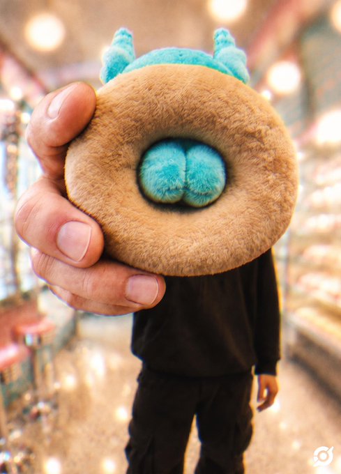 A hand holds up a small brown furry plush toy with teal horns and a teal patch on its back, showcasing a Koda Cake item available at ApeFest.
