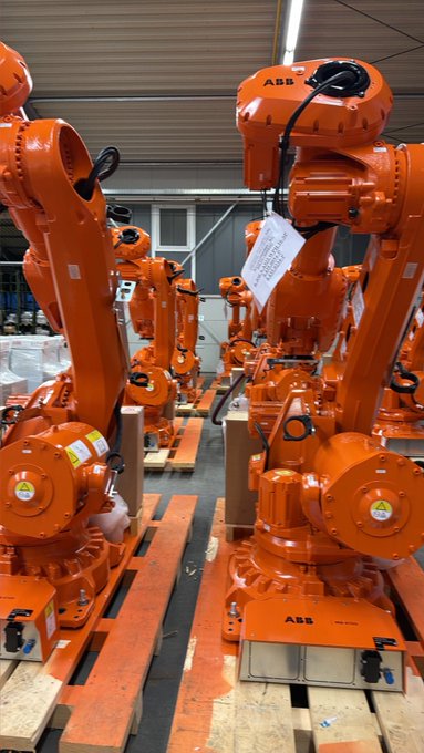 A warehouse or factory floor with multiple orange ABB industrial robotic arms lined up on wooden pallets.