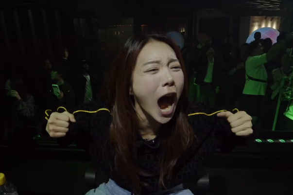 A woman with long brown hair screams with her mouth wide open while raising her fists, appearing excited in a dark venue with other people in the background.