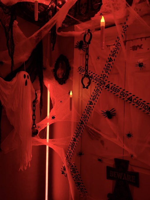 Halloween decoration scene bathed in red light featuring a hanging ghost, cobwebs, fake spiders, chains, lit candles, "BEWARE" sign, and yellow "CAUTION KEEP OUT" tape.