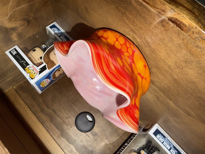 A handblown glass vase with vibrant red, orange, and yellow murrine pattern and a pink interior sits on a wooden surface next to Funko POP! boxes, one labeled 'AT-AT DRIVER' and another 'FUNKO DERPY'.
