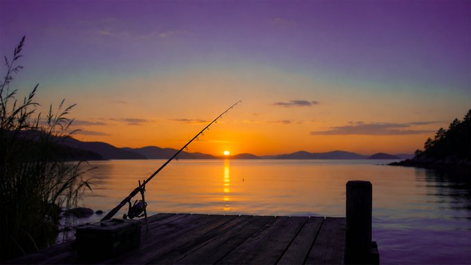 A serene sunset photo over a calm body of water, with a fishing rod set up on a wooden dock and mountains silhouetted in the distance.