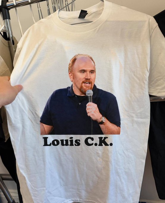 A white t-shirt hanging on a rack features a printed photo of comedian Louis C.K. holding a microphone, with his name printed below.